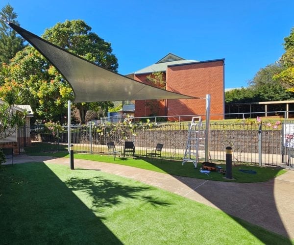 A dark grey triangular shade sail is installed over a grassy lawn and a paved walkway.