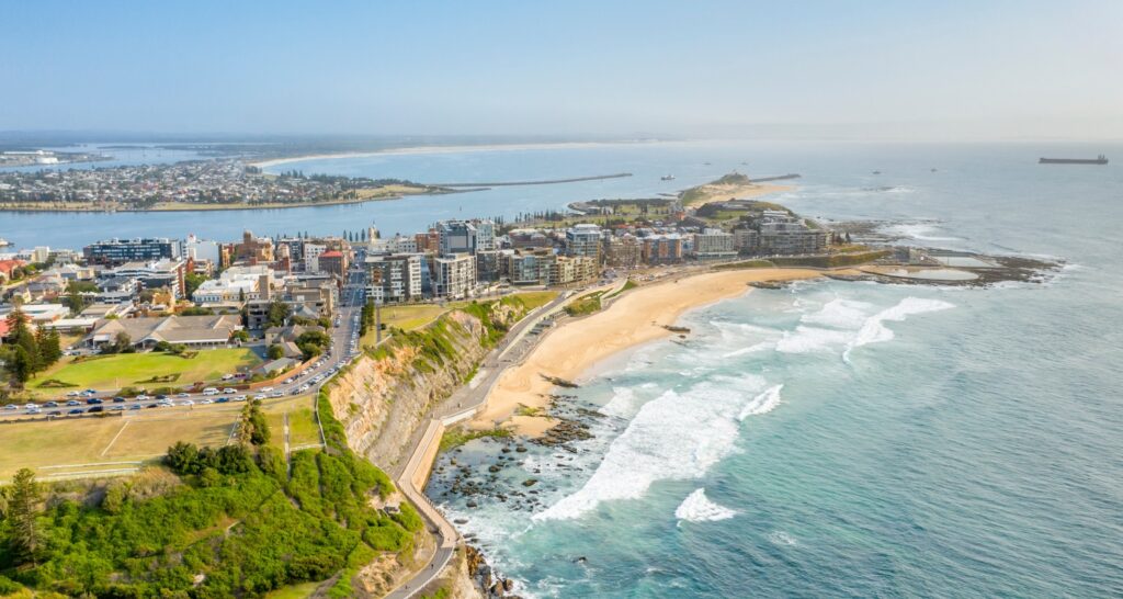 An aerial panorama of a coastal city featuring a long, sandy beach and rocky shoreline along a bright blue ocean.