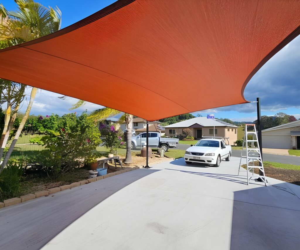 A vibrant orange shade sail provides cover over a white concrete driveway.