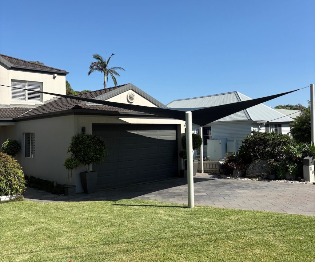 A dark grey triangular shade sail is tensioned over a paved driveway in front of a modern home with a grey garage door.