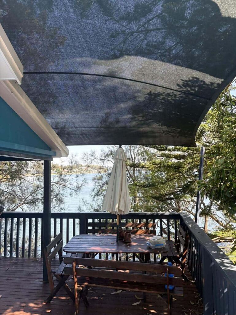 A dark rectangular shade sail is installed over a wooden deck with a picnic table and a white umbrella.