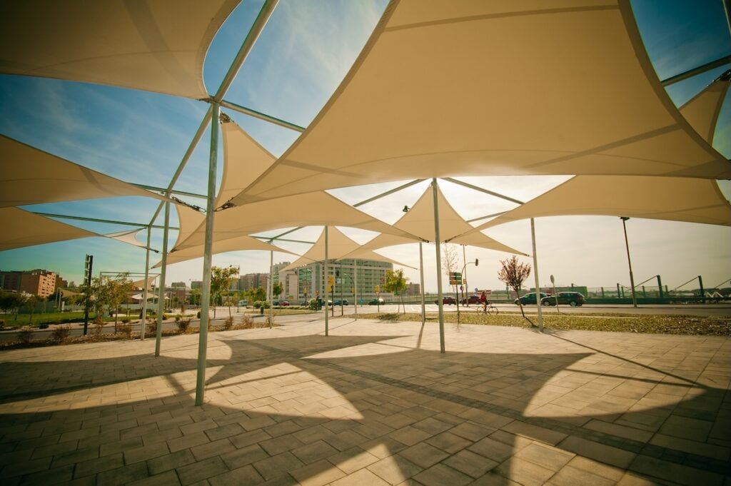 A network of numerous triangular cream shade sails are connected to tall metal poles to cover a large, paved public plaza.