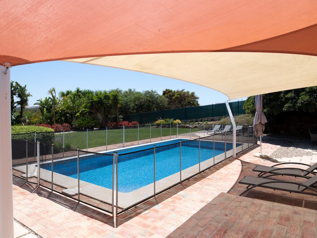 fenced swimming pool in garden with sunbeds under a shade sail