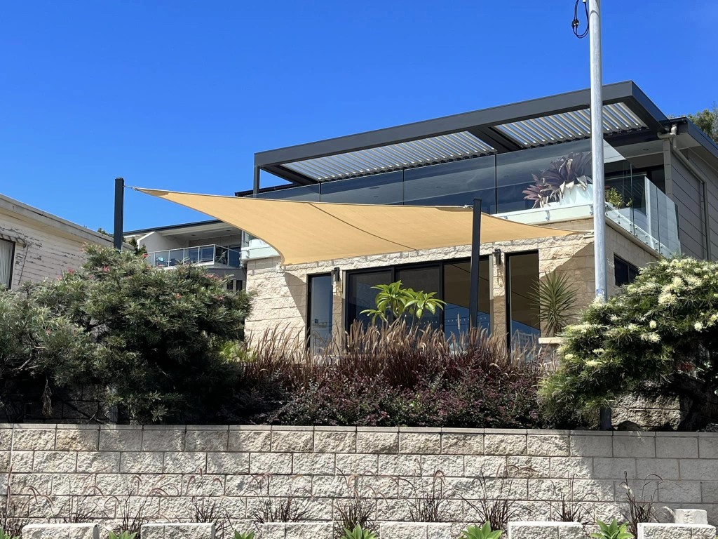 A triangular cream shade sail is installed over a stone patio of a multi-storey modern home.